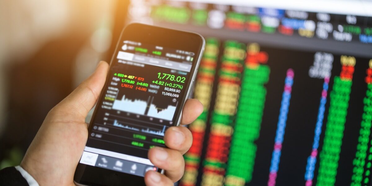 A point of view of looking at a smartphone with online trading information in front of a desktop with more trading info.