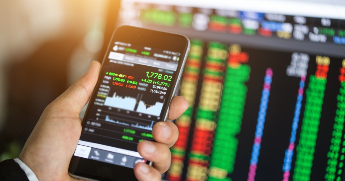 A point of view of looking at a smartphone with online trading information in front of a desktop with more trading info.