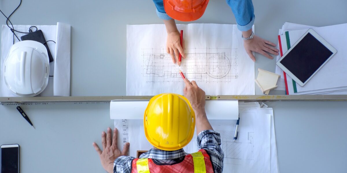 An overhead view of two people in hard hats and safety vests leaning over a desk with pencils and blueprints.