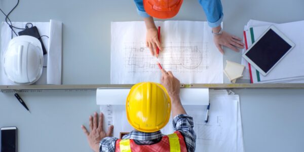 An overhead view of two people in hard hats and safety vests leaning over a desk with pencils and blueprints.