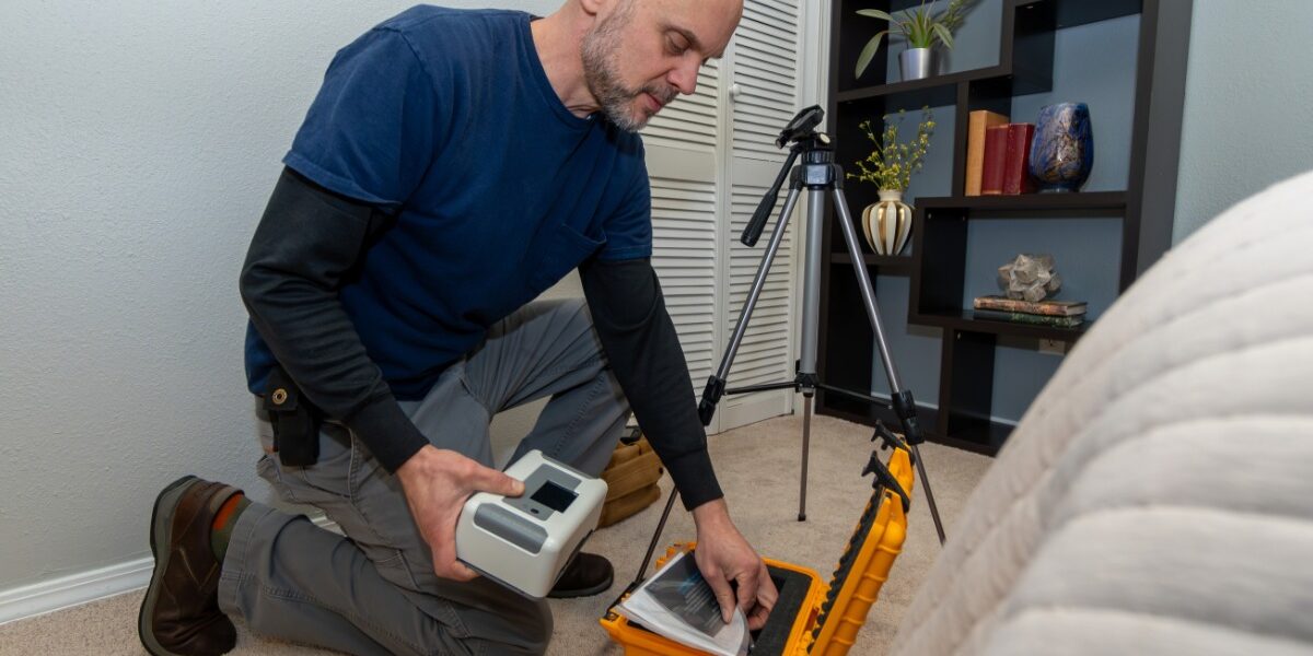 A man wearing a blue T-shirt and dark jeans is holding a small machine while kneeling and looking through a toolbox.