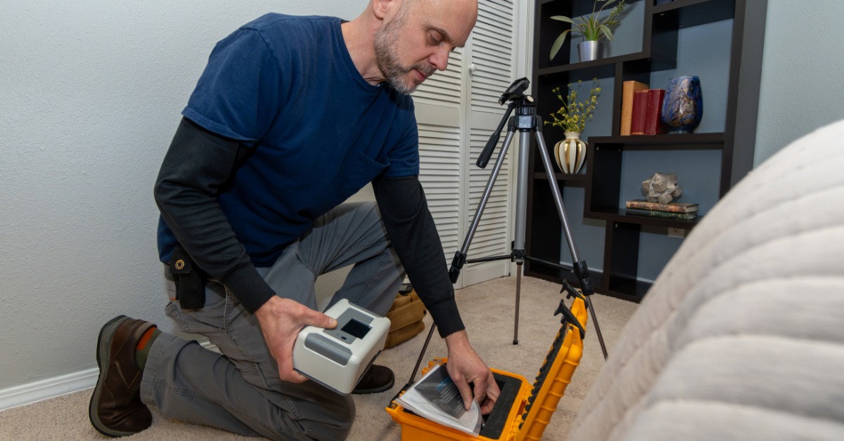 A man wearing a blue T-shirt and dark jeans is holding a small machine while kneeling and looking through a toolbox.