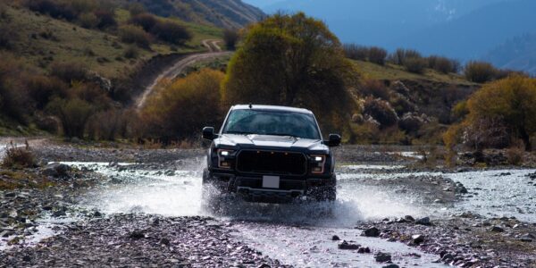 A pickup truck is driving through a puddle of water on a rocky surface. Mountains are in the distance.