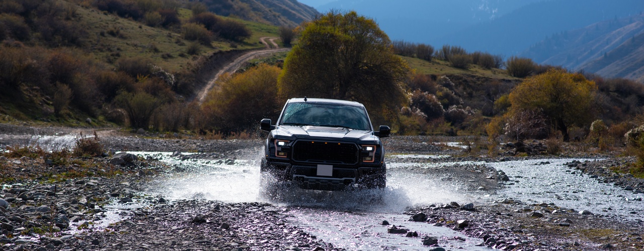 A pickup truck is driving through a puddle of water on a rocky surface. Mountains are in the distance.