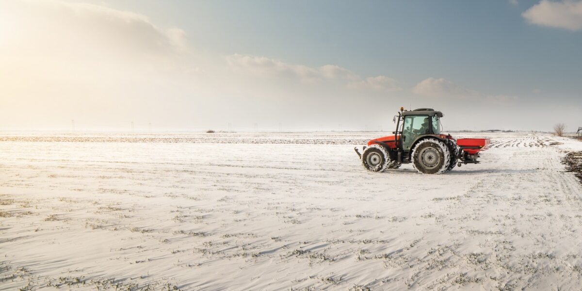 A red tractor is driving on farmland covered in a thin layer of snow. The rising sun brightens the field.