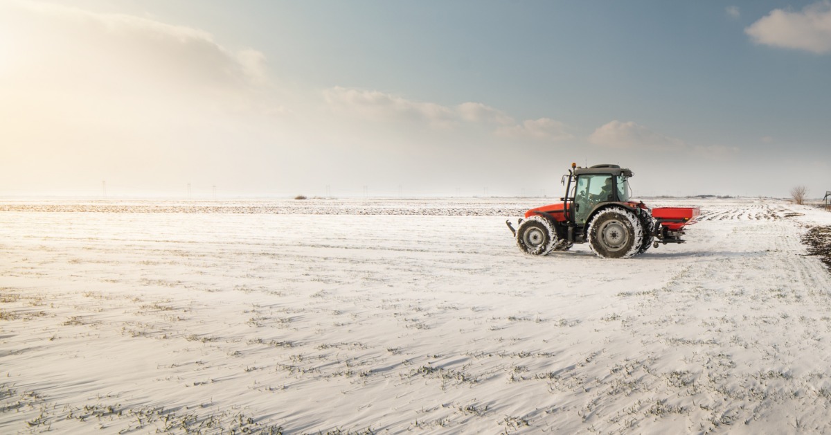 A red tractor is driving on farmland covered in a thin layer of snow. The rising sun brightens the field.