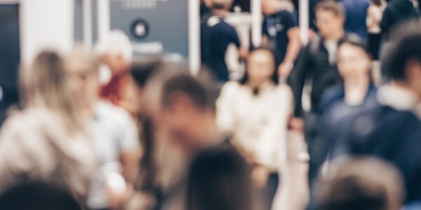 A blurred crowd walking through an exhibition hall with trade show booths, signs, ceiling lights, and open walkways.