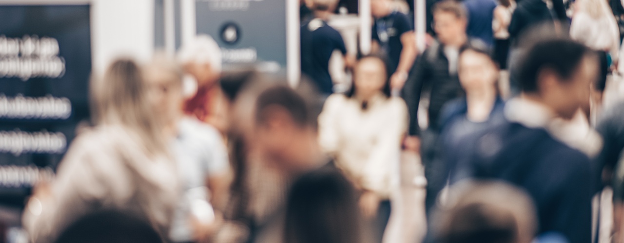 A blurred crowd walking through an exhibition hall with trade show booths, signs, ceiling lights, and open walkways.
