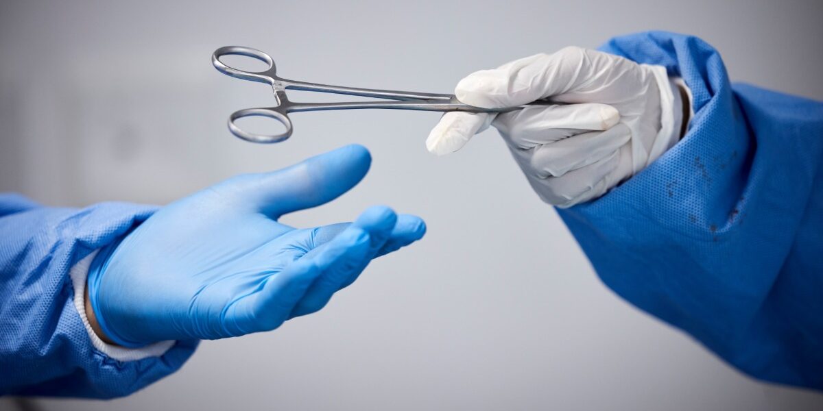 One medical professional wearing white gloves is handing another professional a pair of medical scissors.