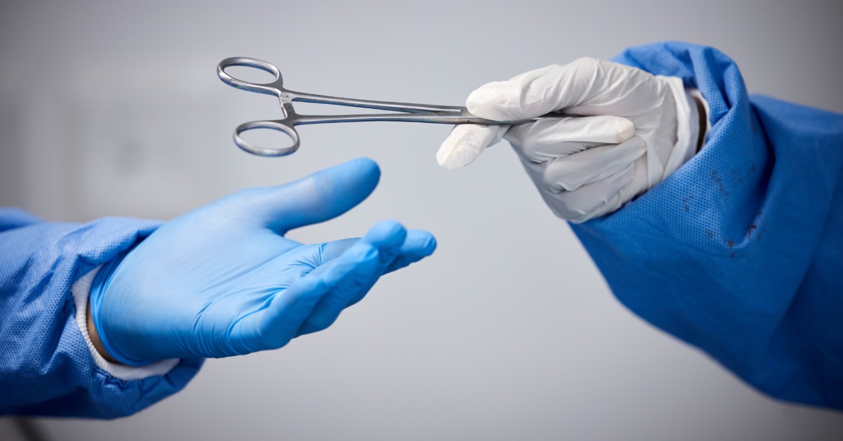 One medical professional wearing white gloves is handing another professional a pair of medical scissors.
