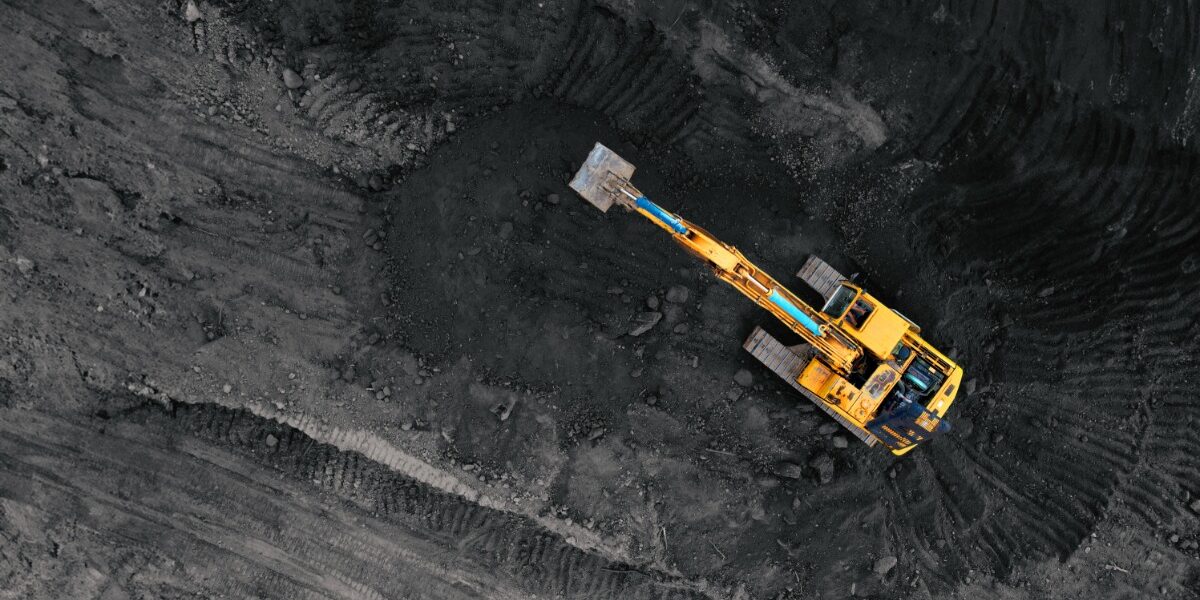 Aerial view of a yellow excavator on dark, churned soil, surrounded by sweeping tire tracks and rough terrain.