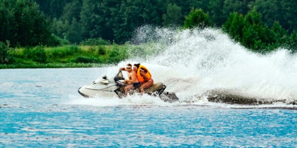 A man and a woman are riding on a white jet ski and creating large waves. The woman is wearing an orange life vest.