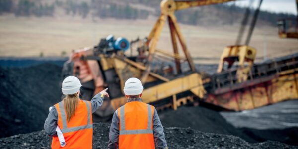 Two workers wearing safety vests and white helmets looking at a large crane in a mining site between mountains of coal.