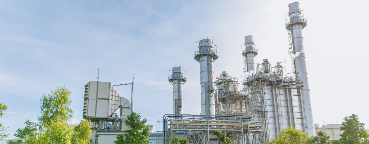An industrial gas processing facility with tall metal towers, storage tanks, and pipelines surrounded by green trees.