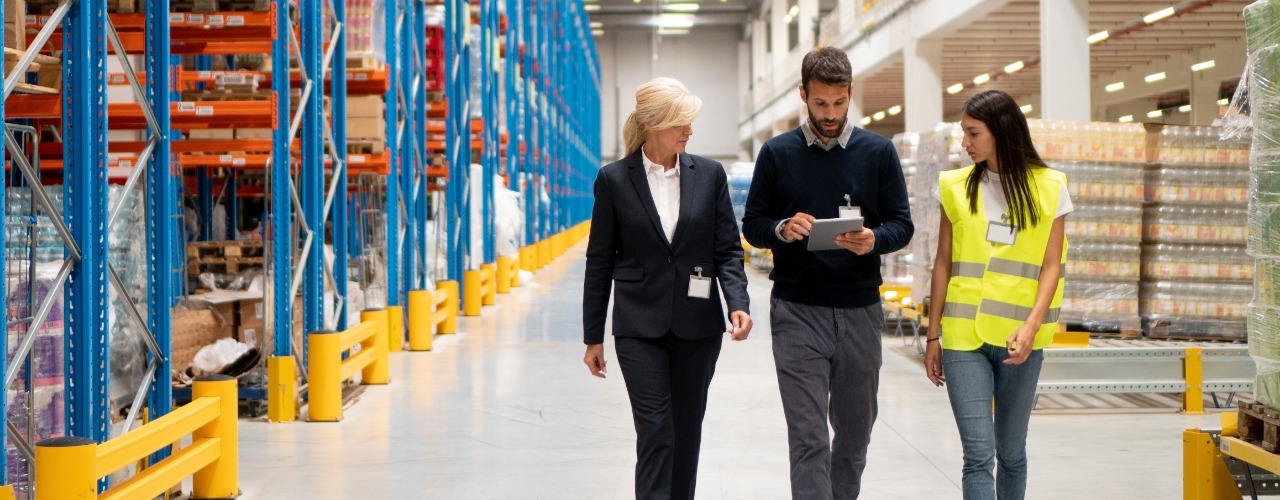 Three business professionals walk through a warehouse aisle as they audit the business's efficiency demands.