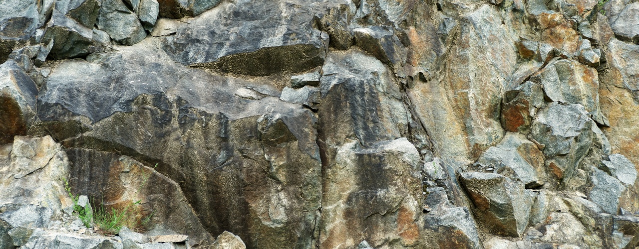 Close-up of a rugged rock face with jagged gray and brown stone formations, deep cracks, and scattered loose rocks.
