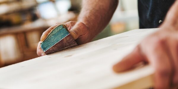 A close-up of the hands of a man using a sanding block to smooth the side of a flat wooden beam on a base.
