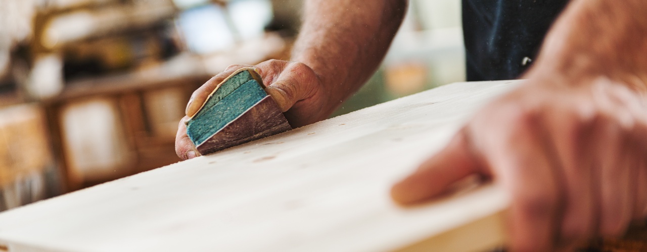 A close-up of the hands of a man using a sanding block to smooth the side of a flat wooden beam on a base.
