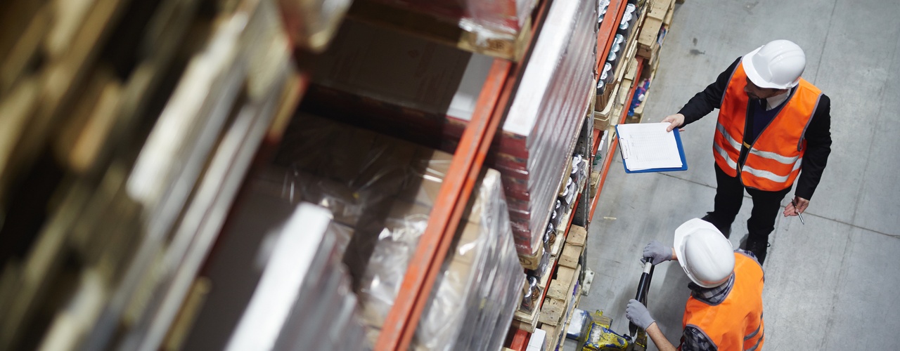 An aerial view of two workers wearing safety vests and white helmets using a pallet jack inside a large warehouse.