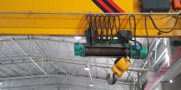 A close-up view shows a yellow overhead crane in a large metal building. The crane hook is all the way up.