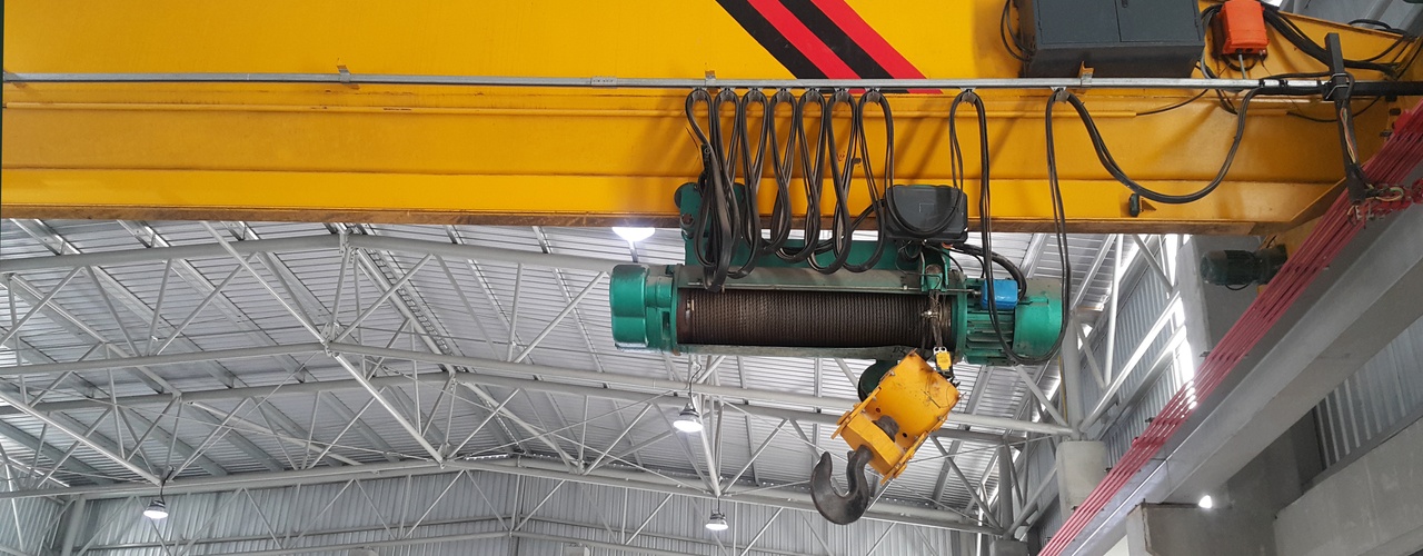 A close-up view shows a yellow overhead crane in a large metal building. The crane hook is all the way up.