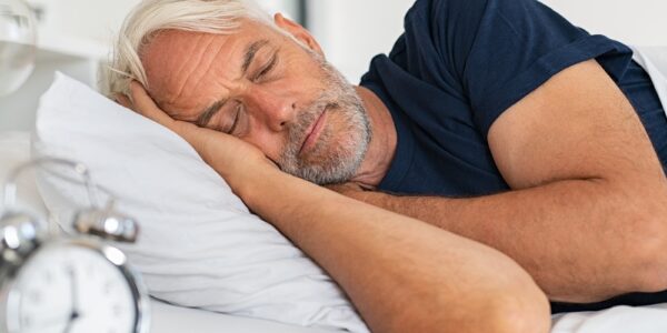 A senior man wearing a blue shirt sleeping on his right side next to a nightstand with a metal alarm clock.