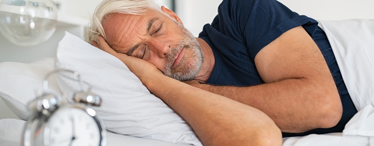 A senior man wearing a blue shirt sleeping on his right side next to a nightstand with a metal alarm clock.